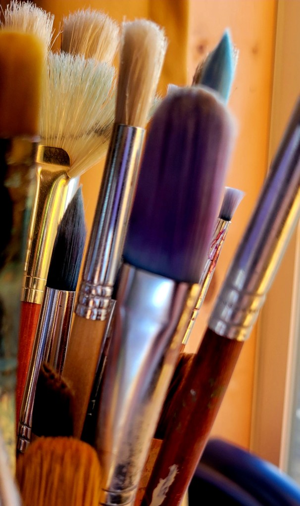 Close-up photograph of well-used paintbrushes with varied bristle shapes and sizes, arranged upright in a container under warm studio lighting.